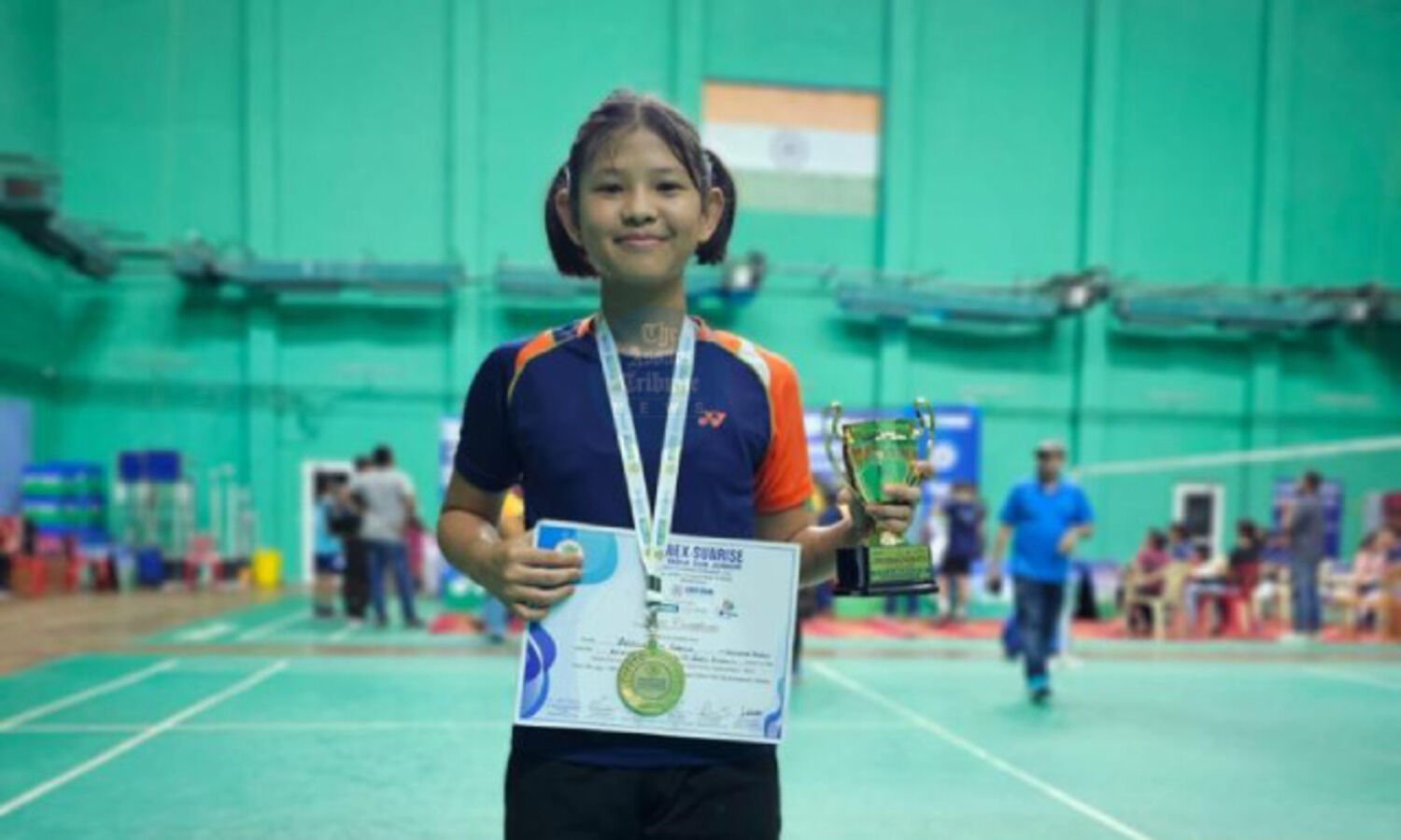 Jesicca Neyi Saring on a badminton court, wearing her gold medal and holding her Yonex-Sunrise certificate and a trophy